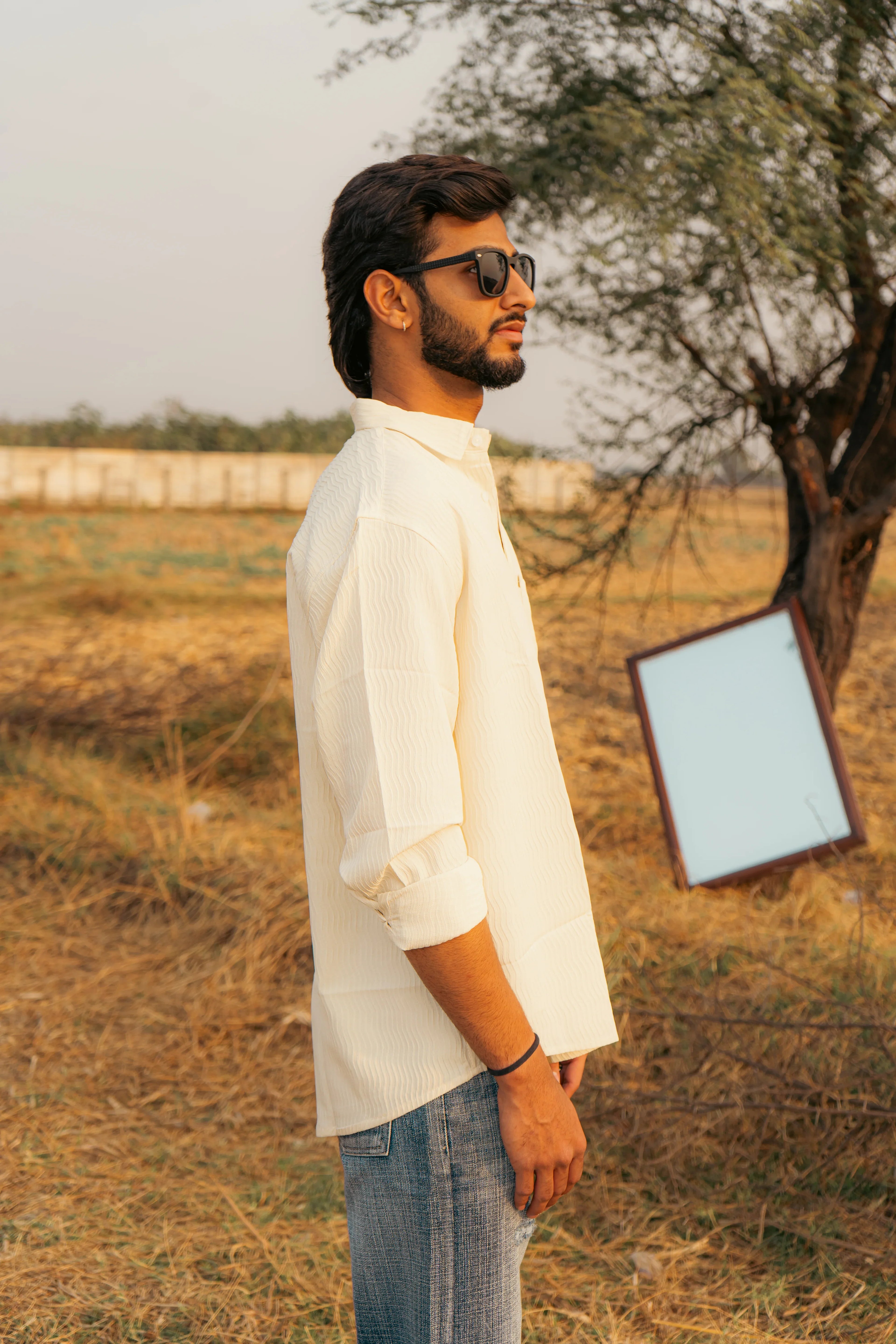 Man in textured off-white shirt and jeans, wearing sunglasses, standing outdoors near a tree