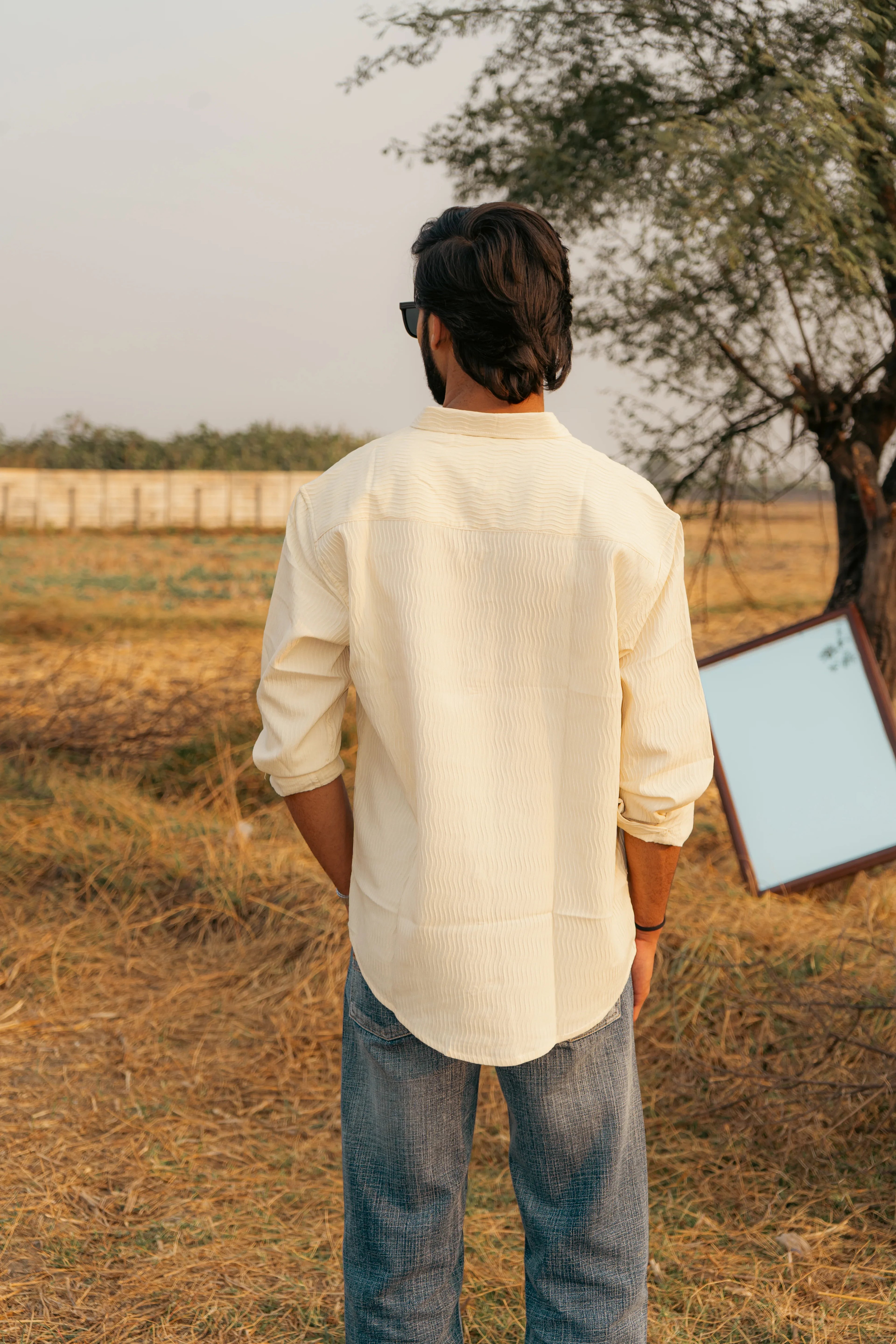 Man in cream textured shirt and jeans standing outdoors by a tree and mirror