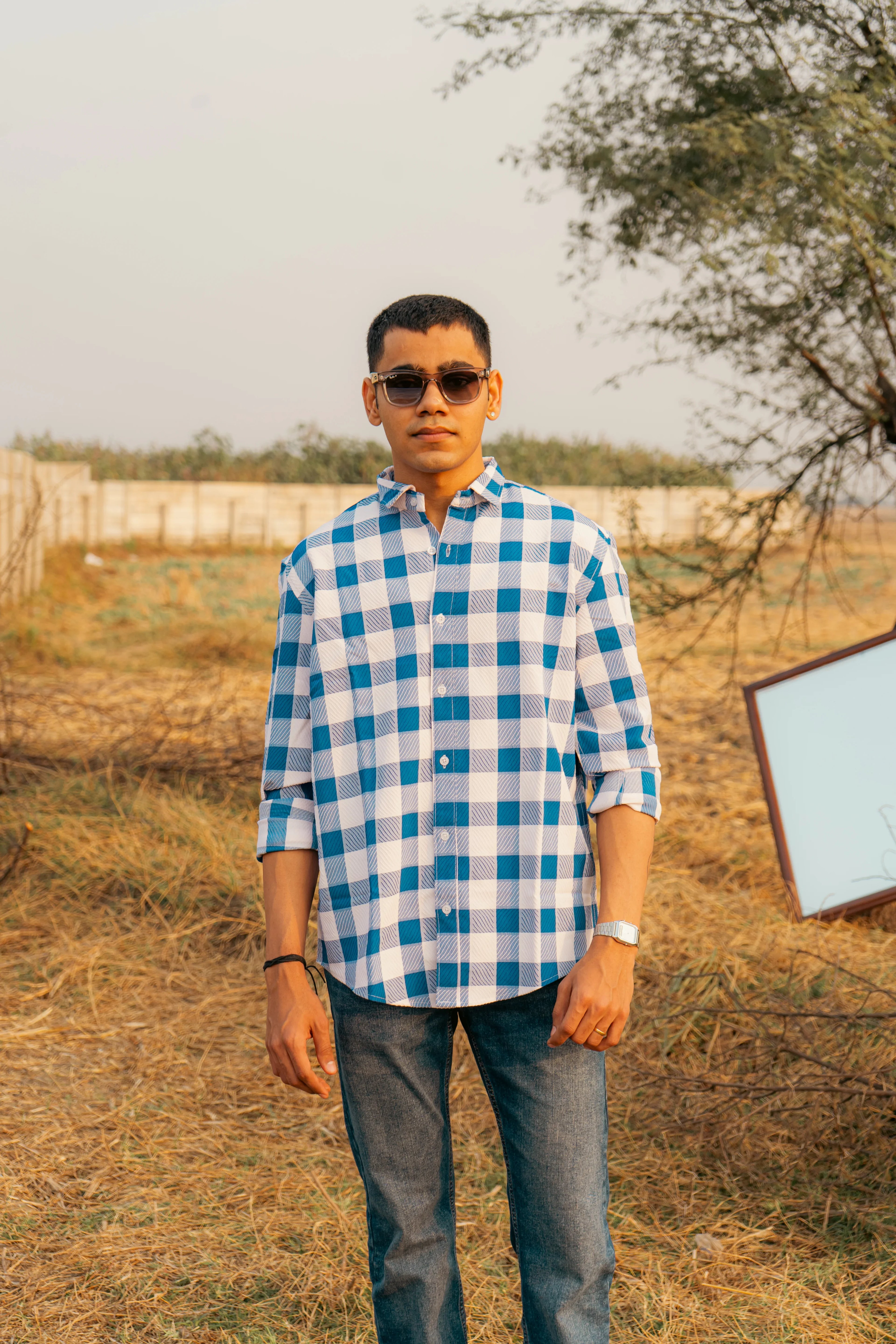 Man outdoors wearing blue and white plaid shirt, jeans, and sunglasses in natural light