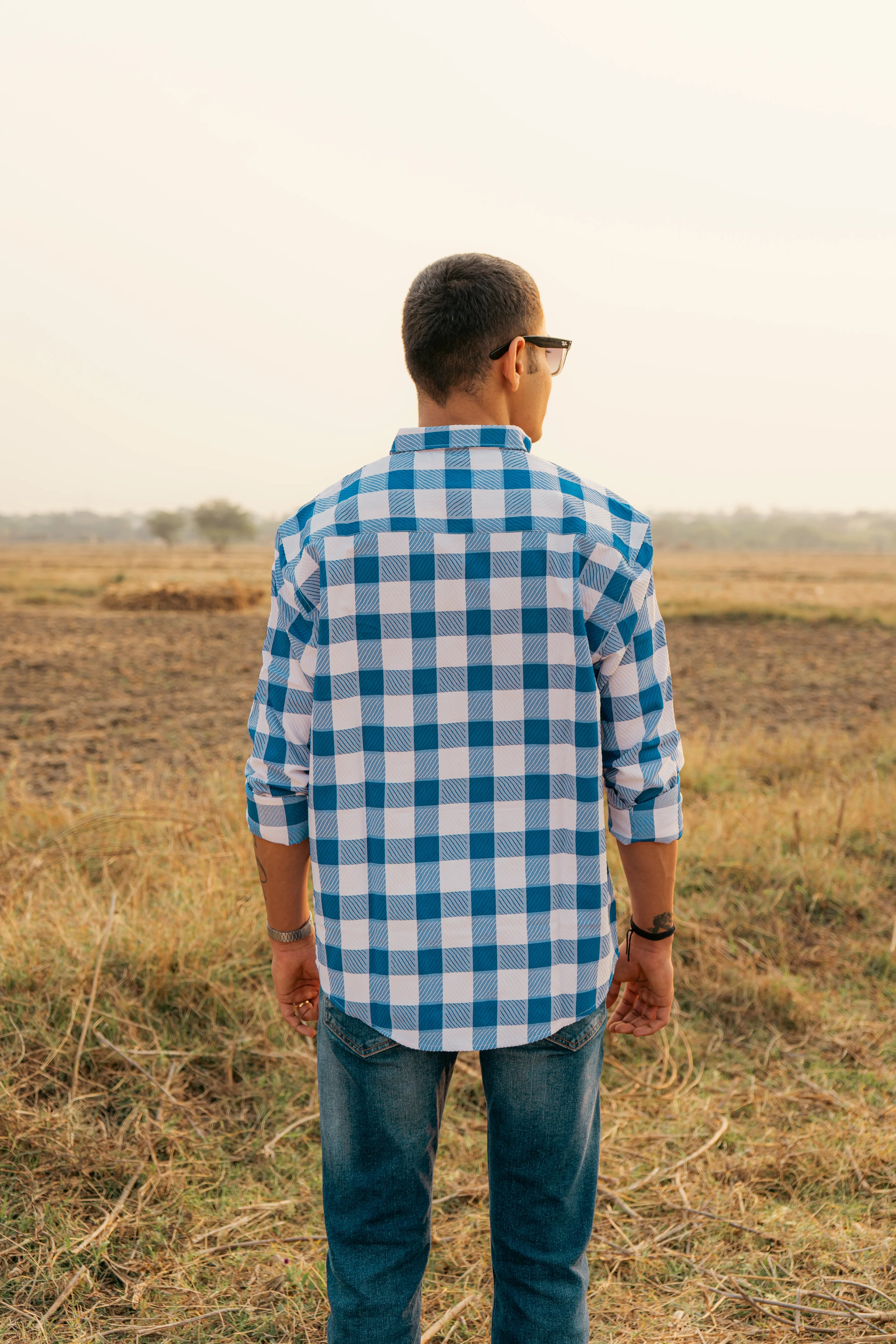 Man wearing blue and white checkered shirt and jeans standing in a field, back view
