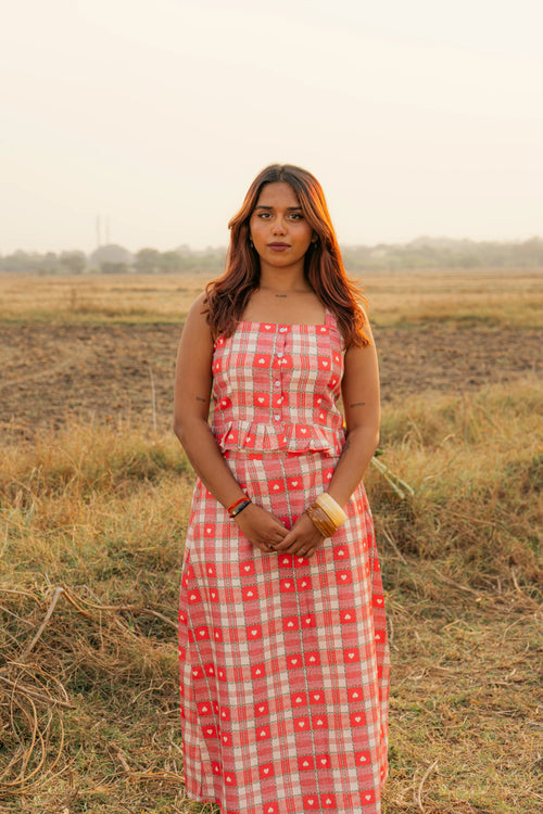 Woman in red plaid sleeveless dress with white heart pattern standing in a grassy field at sunset