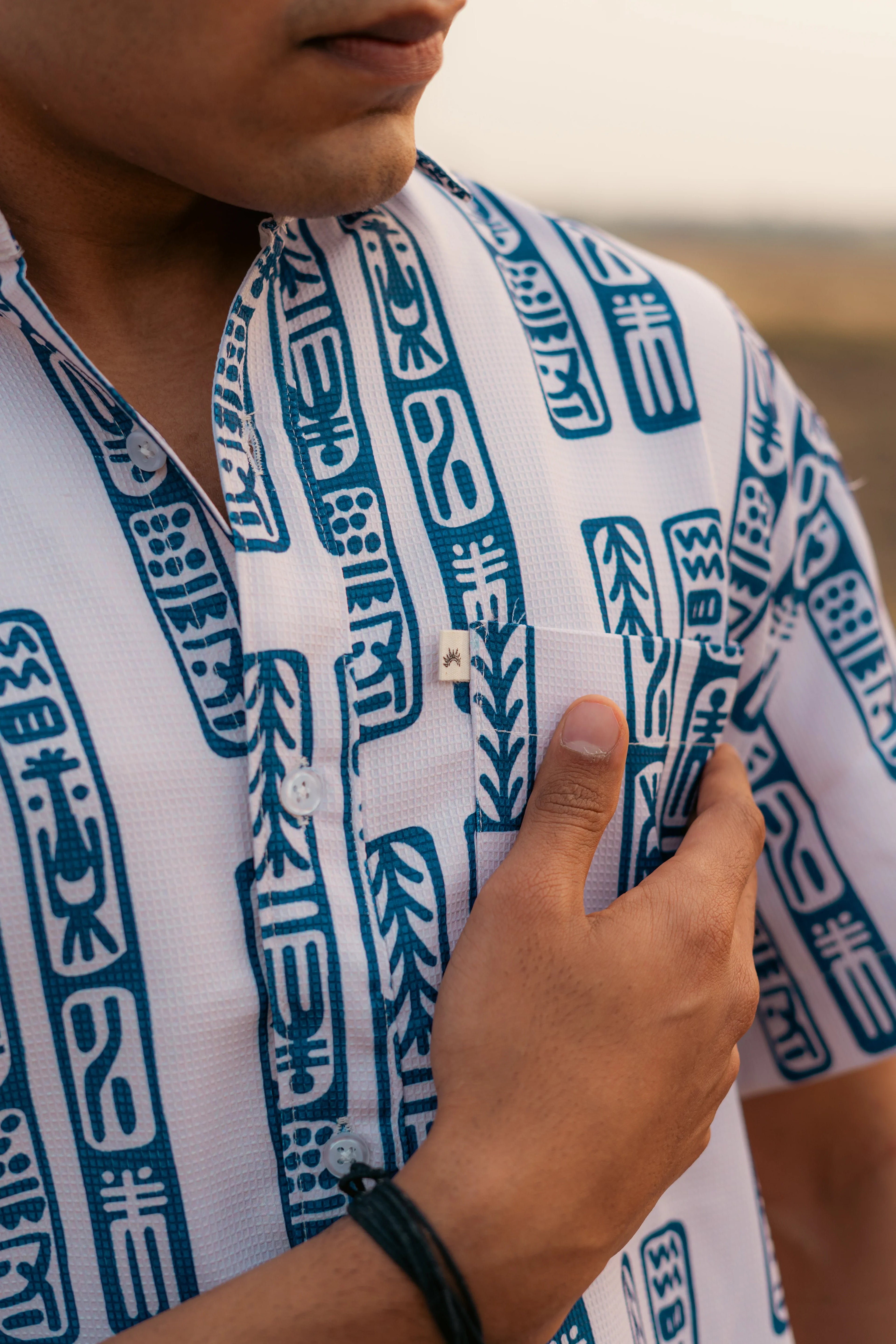 Man wearing white shirt with blue geometric print, hand touching chest pocket outdoors