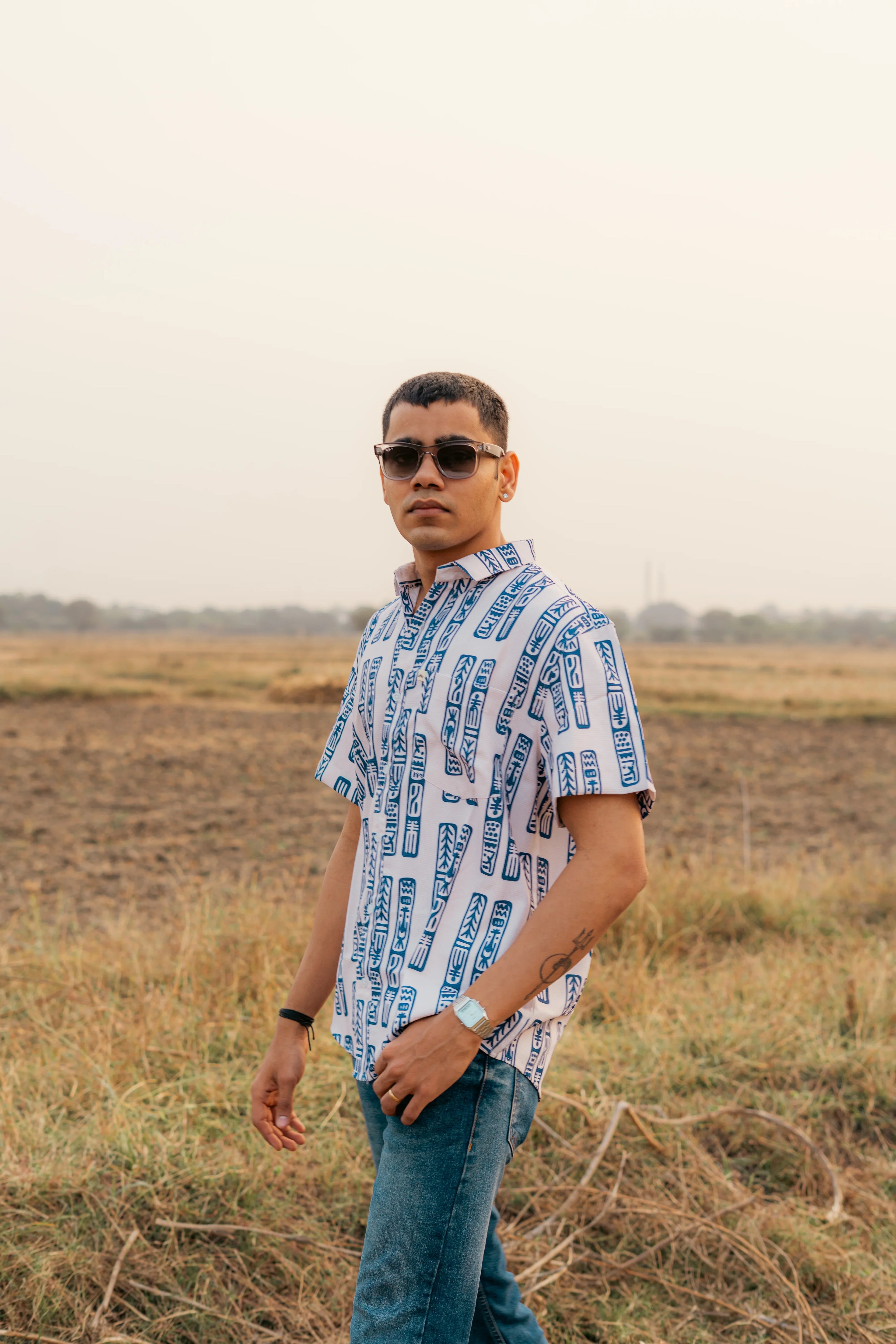 Man in sunglasses wearing patterned short sleeve shirt and jeans standing outdoors