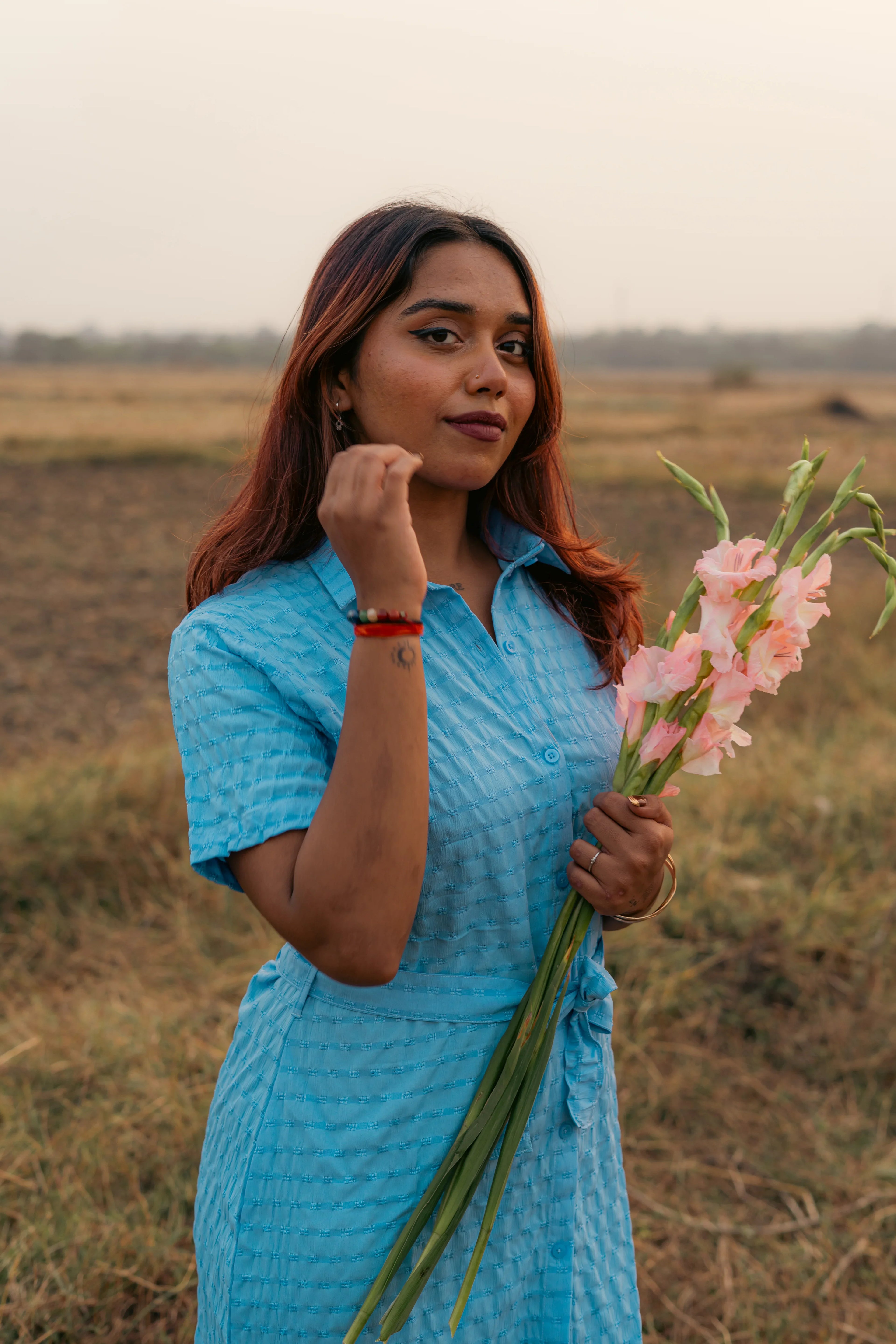Woman in a blue textured dress holding pink flowers in an open field at sunset