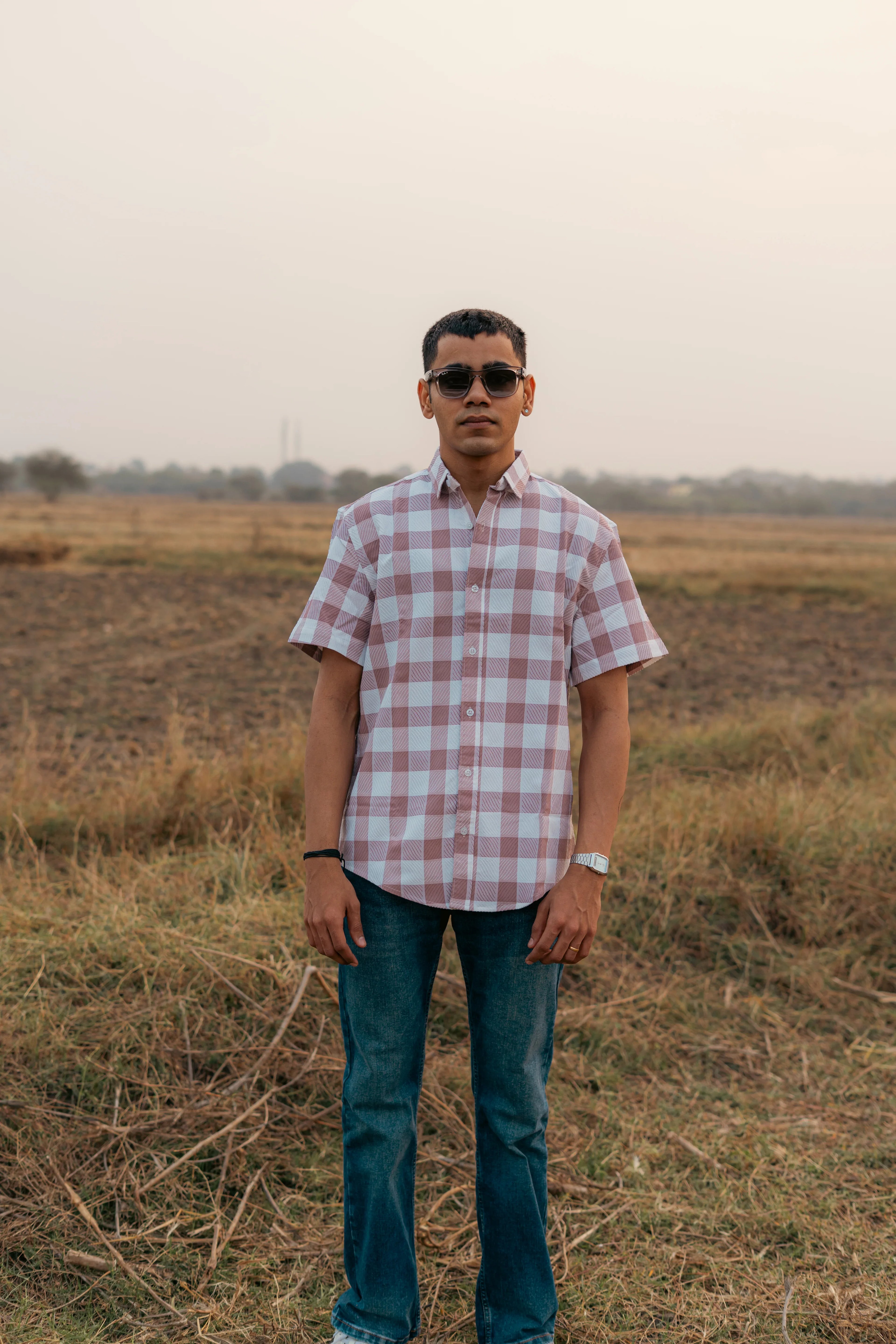 Man wearing sunglasses, pink plaid short sleeve shirt, and jeans standing outdoors