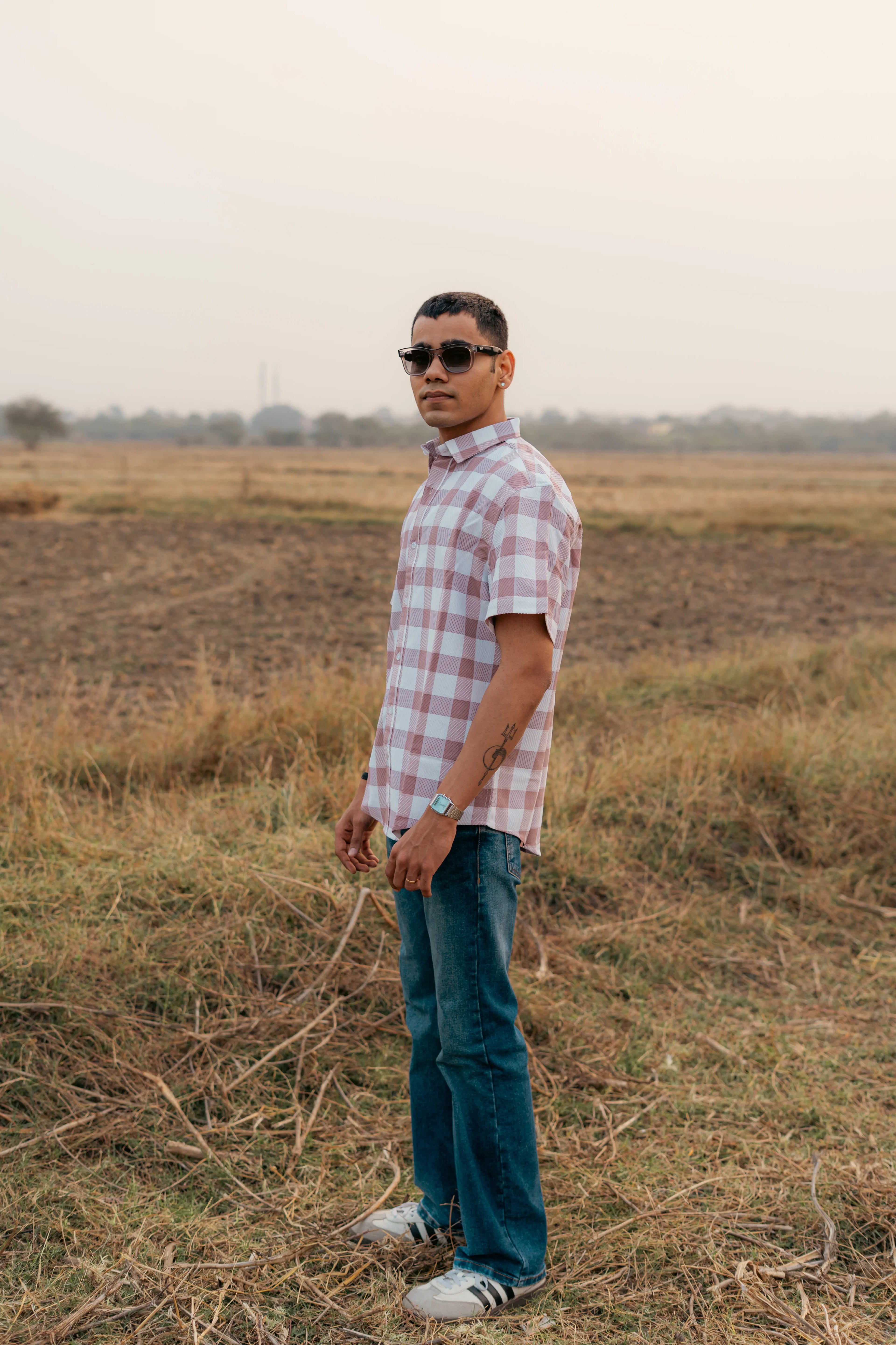 Man in a short-sleeve plaid shirt, jeans, and sunglasses standing in a field