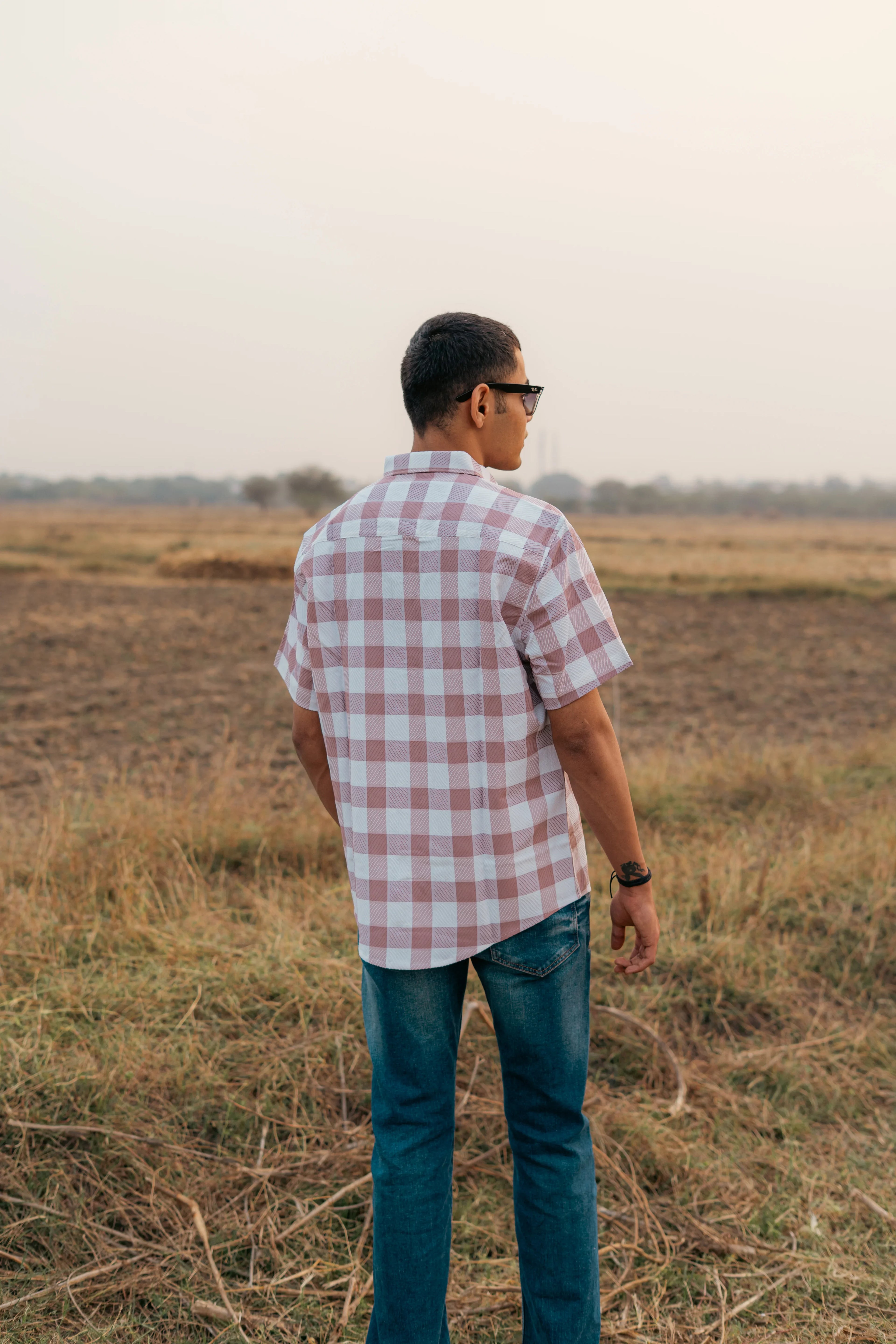 Man wearing short sleeve plaid shirt and jeans standing in a field at sunset