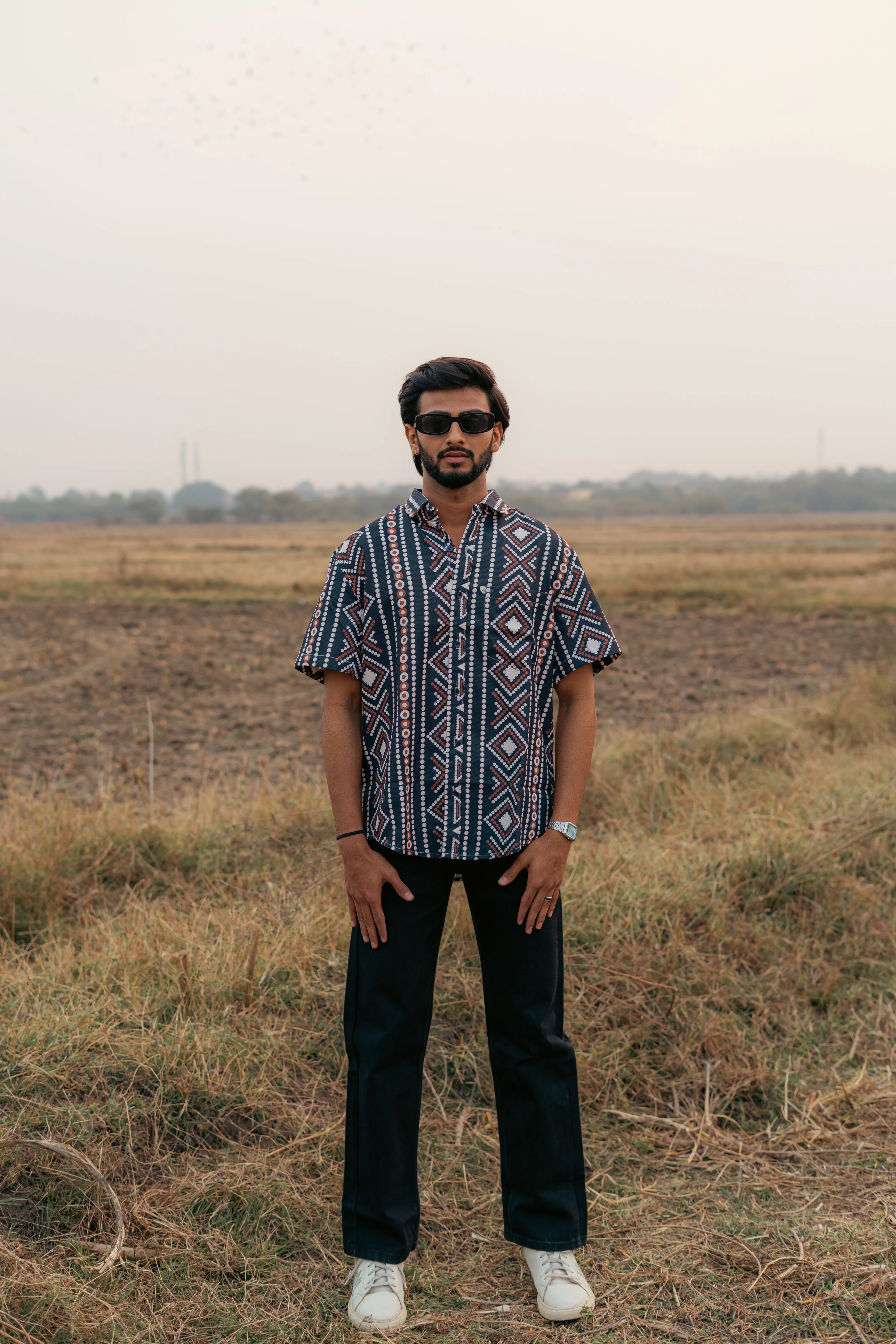 Man in geometric print short sleeve shirt, black pants, and sunglasses standing in a field