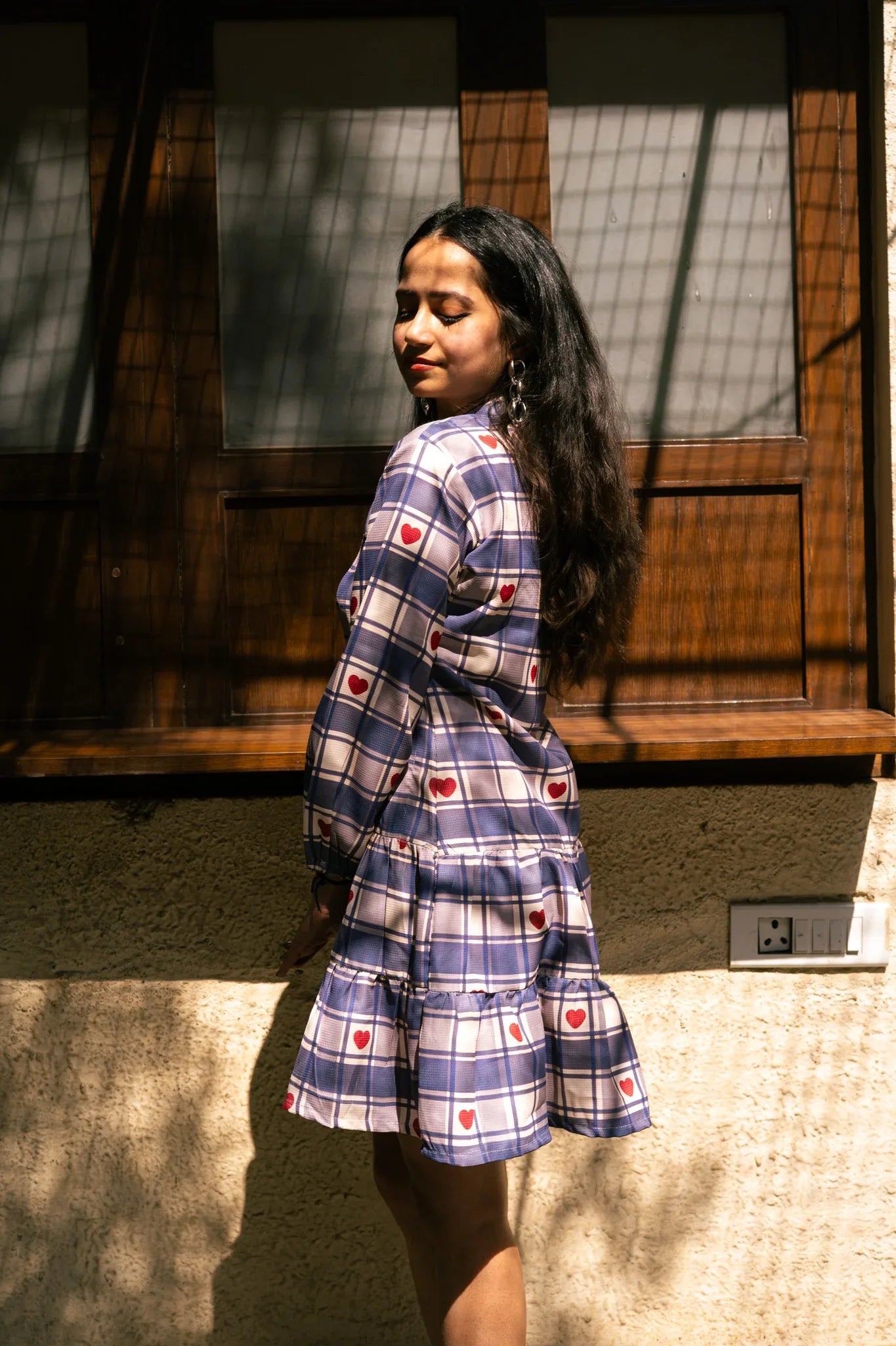 Woman in a blue plaid dress with red heart prints standing by a sunlit window