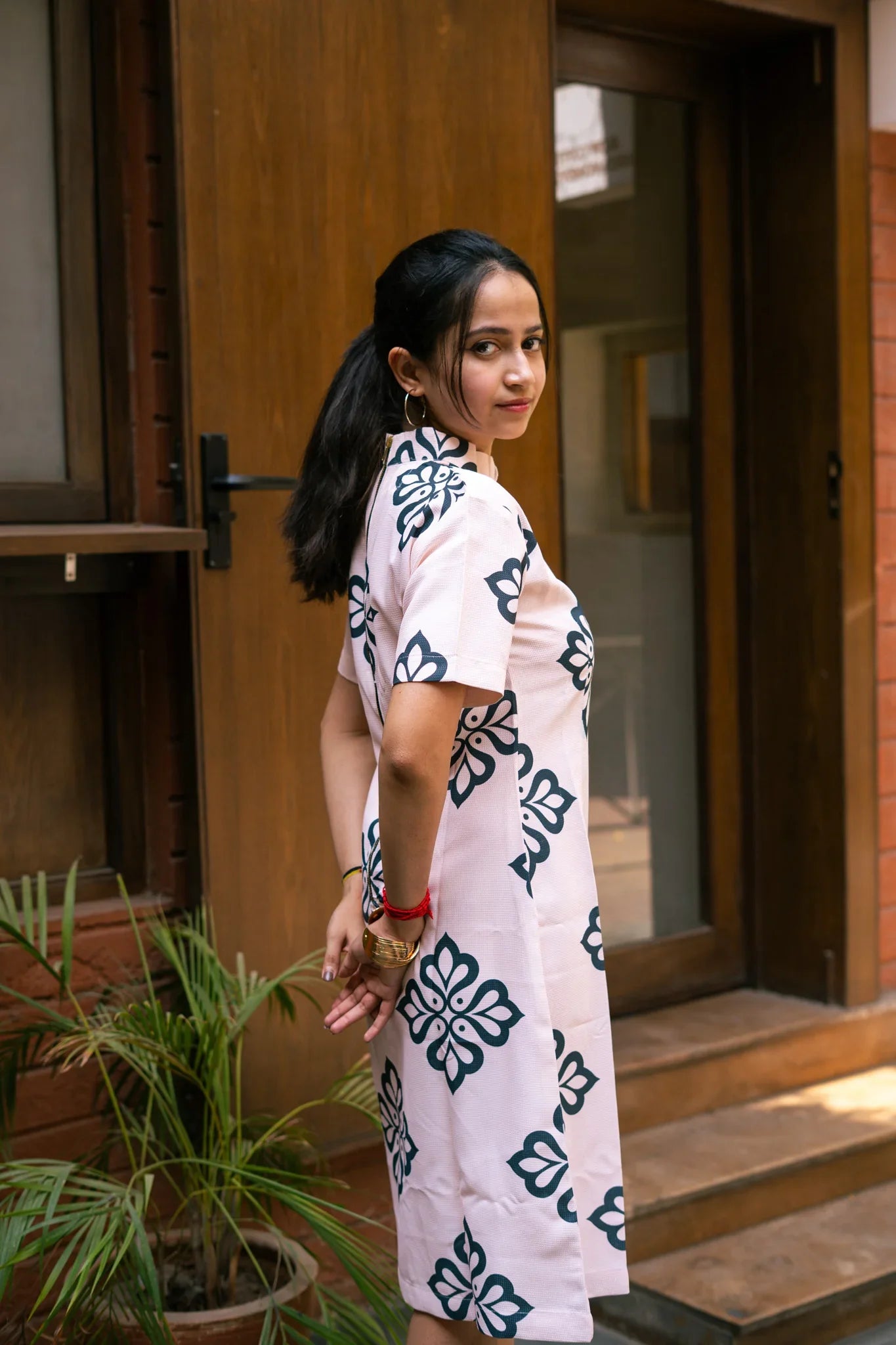 Woman in a light pink dress with bold floral prints standing outdoors by a wooden door